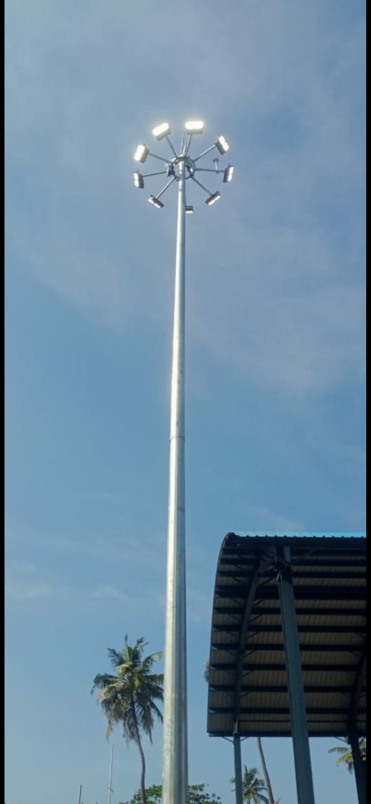 A high-mast lighting pole with multiple floodlights, raised and lit against a clear sky